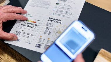 Person hält einen digitalen Gästepass und liest ein gedrucktes Dokument bei einem Tisch, eine Person fotografiert es mit einem Smartphone. | © Kleinwalsertal Tourismus eGen • Fotograf: Oliver Farys