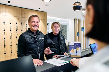 Zwei lachende Personen am Empfang bei einer Registrierung im Winter 2025 im Kleinwalsertal, betreut von einem Mitarbeiter. | © Kleinwalsertal Tourismus eGen • Fotograf: Oliver Farys