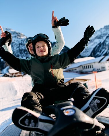 Kids sledging in Kleinwalsertal, smiling happily, with mountains in the background, winter scene. | © Kleinwalsertal Tourismus eGen • Photographer: Oliver Farys