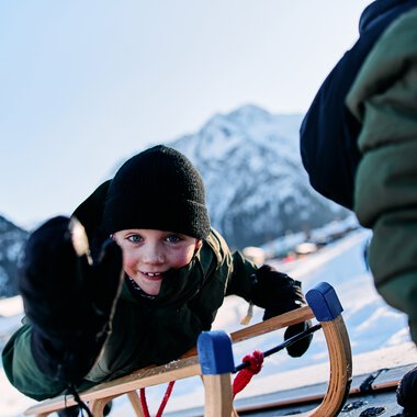 Kind beim Rodeln im Kleinwalsertal, lachend und in Wintersportkleidung, mit Bergen im Hintergrund. | © Kleinwalsertal Tourismus eGen • Fotograf: Oliver Farys