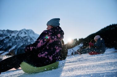 Children sledding in the mountains of Kleinwalsertal on a sunny winter day, with snow-covered landscape and mountain peaks in the background. | © Kleinwalsertal Tourismus eGen • Photographer: Oliver Farys