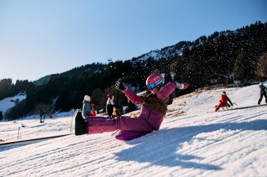 Children sledding in winter at Kleinwalsertal, laughing and playing on a snowy slope surrounded by mountains. | © Kleinwalsertal Tourismus eGen • Photographer: Oliver Farys