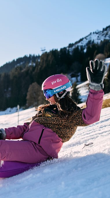 A girl sliding down a snowy slope, smiling and waving, in Kleinwalsertal with mountains in the background, enjoying winter snow tubing with other children around. | © Kleinwalsertal Tourismus eGen • Photographer: Oliver Farys