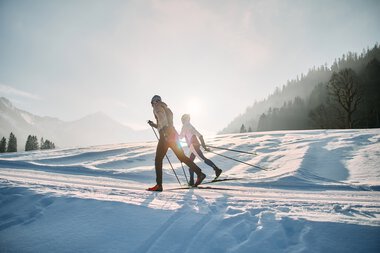 Zwei Skifahrer beim klassischen Langlauf auf der Schwarzwasserloipe im Kleinwalsertal bei Sonnenaufgang, umgeben von verschneiter Landschaft und Bergen. | © Kleinwalsertal Tourismus eGen • Fotograf: Oliver Farys