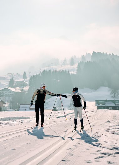 Two cross-country skiers on the winter trail in Kleinwalsertal, with snow-covered houses and mountains in the background, cloudy sky, winter landscape. | © Kleinwalsertal Tourismus eGen • Photographer: Oliver Farys