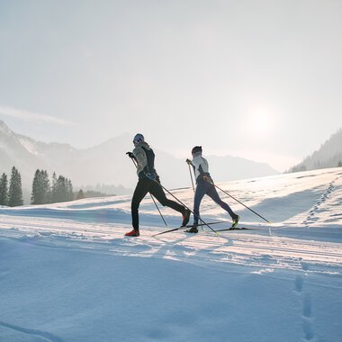 Zwei Skifahrer ziehen im frühen Morgenlicht durch die verschneite Schwarzwasserloipe im Kleinwalsertal, umgeben von Bergen und Tannen | © Kleinwalsertal Tourismus eGen • Fotograf: Oliver Farys