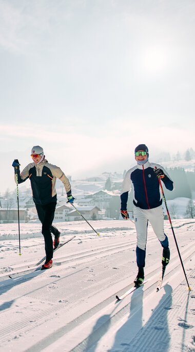 Two cross-country skiers ski on a snowy trail in Kleinwalsertal with mountains in the background. | © Kleinwalsertal Tourismus eGen • Photographer: Oliver Farys