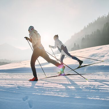 Two people cross-country skiing on the Schwarzwasserloipe in Kleinwalsertal, surrounded by snow-covered scenery and mountains in the background during sunrise. | © Kleinwalsertal Tourismus eGen • Photographer: Oliver Farys