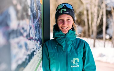 A smiling woman in winter gear with ski goggles stands next to an information sign in the snow, with trees and a snowy landscape in the background. | © Kleinwalsertal Tourismus eGen • Photographer: Oliver Farys