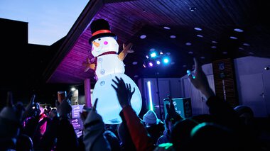 Großer aufblasbarer Schneemann auf einer Bühne bei einer Veranstaltung im Kleinwalsertal, mit Publikum und bunten Lichtern. | © Kleinwalsertal Tourismus eGen • Fotograf: Oliver Farys