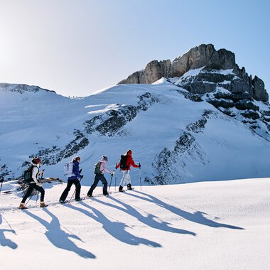 Six people snowshoeing in snowy mountain landscape in Kleinwalsertal, with sunshine in the sky. | © Kleinwalsertal Tourismus eGen • Photographer: Oliver Farys