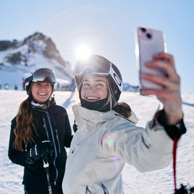 Two happy skiers on Kanzelwand in Kleinwalsertal, one taking a selfie in front of snowy mountains under the sunshine. | © Kleinwalsertal Tourismus eGen • Photographer: Oliver Farys