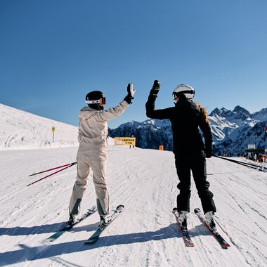 Two skiers giving a high-five on a snowy mountain slope, with the Alps in the background and clear blue sky. | © Kleinwalsertal Tourismus eGen • Photographer: Oliver Farys