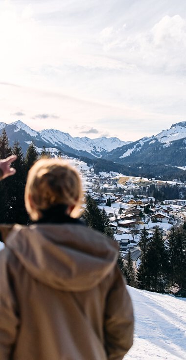 Two people overlook a snowy mountain and valley landscape with houses, forests, and mountains in the background from a snow-covered hill. | © Kleinwalsertal Tourismus eGen • Photographer: Oliver Farys