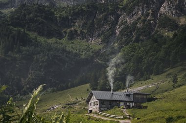 Landscape with green hills, a small cabin with smoke coming from the chimneys, surrounded by mountains and clouds in the sky. | © Kleinwalsertal Tourismus eGen • Photographer: Robert Niederwolfsgruber