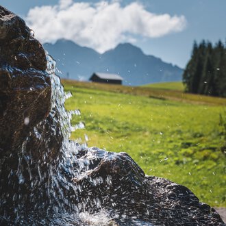 Close-up of a waterfall or flowing creek in a green, mountainous landscape with clouds and a small house in the background. | © Kleinwalsertal Tourismus eGen • Photographer: Robert Niederwolfsgruber