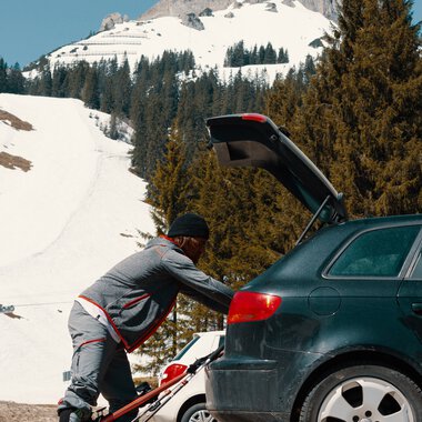 Mann beim Skituning neben einem Auto im Winteralpenland, mit schneebedecktem Berg im Hintergrund. | © Kleinwalsertal Tourismus eGen • Fotograf: Distillery