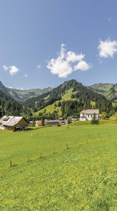 Photo of a green hillside with houses and mountains under a blue sky with clouds. | © Kleinwalsertal Tourismus eGen • Photographer: Steffen Berschin 