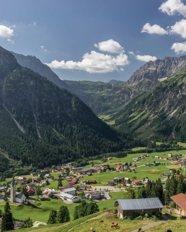 Panoramaaufnahme des Bergdorfes Mittelberg in den Alpen mit grünen Hügeln, Bergen im Hintergrund und einer blauen wolkenverhangenen Himmel. | © Kleinwalsertal Tourismus eGen • Fotograf: Steffen Berschin 