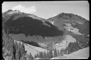 Black and white photo of mountain landscape with cabins in a valley surrounded by forests, with tall mountains in the background. | © Kleinwalsertal Tourismus eGen • Karl Max Kessler Archiv
