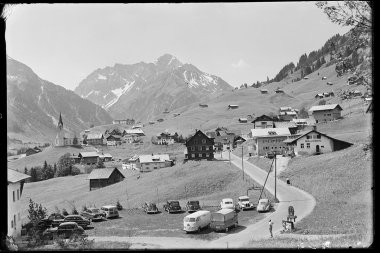 Historisches Schwarz-Weiß-Foto des Ortszentrums von Hirschegg im Kleinwalsertal, mit Kirche, umliegenden Häusern, Autos und den Bergen Widderstein und Hochkalber im Hintergrund. | © Kleinwalsertal Tourismus eGen • Karl Max Kessler Archiv