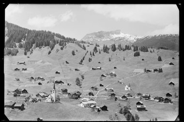 Historische Schwarz-Weiß-Fotoaufnahme von Hirschegg mit verschneiten Ifen, umgeben von Grünflächen, vereinzelten Häusern und einer Kirche. | © Kleinwalsertal Tourismus eGen • Karl Max Kessler Archiv