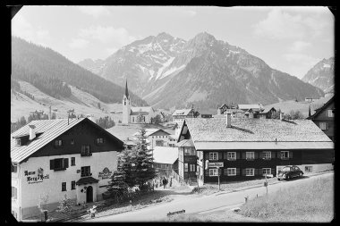 Schwarz-Weiß-Foto eines kleinen österreichischen Dorfes vor majestätischen Bergen, mit einer Kirche, traditionellen Häusern und Autos auf der Straße. | © Kleinwalsertal Tourismus eGen • Karl Max Kessler Archiv