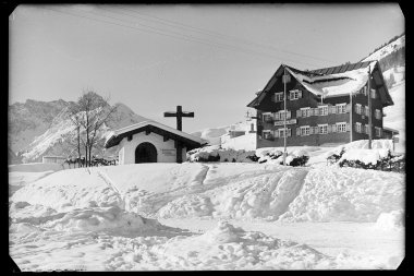 Schnee bedeckter Ort mit Kapelle und Haus in den Bergen, im Winter, historische Fotografie. | © Kleinwalsertal Tourismus eGen • Karl Max Kessler Archiv