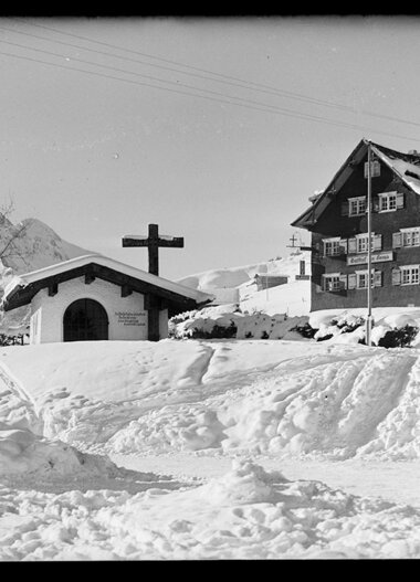 Schnee bedeckter Ort mit Kapelle und Haus in den Bergen, im Winter, historische Fotografie. | © Kleinwalsertal Tourismus eGen • Karl Max Kessler Archiv