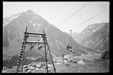 Historisches Foto eines Skiliftantriebs in einem Tal, mit Bergen im Hintergrund und kleinen Gebäuden im Vordergrund. | © Kleinwalsertal Tourismus eGen • Karl Max Kessler Archiv