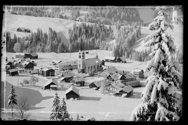 Historisches Foto des verschneiten Riezlern mit Kirche im Zentrum, umgeben von Bergen. | © Kleinwalsertal Tourismus eGen • Karl Max Kessler Archiv