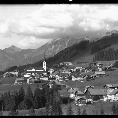 Schwarz-Weiß-Foto des Dorfes Riezlern in den Bergen, mit vielen Häusern, einer Kirche im Zentrum und umliegenden Wiesen und Bergen | © Kleinwalsertal Tourismus eGen • Karl Max Kessler Archiv