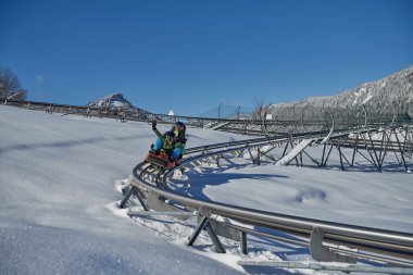 Children on a mountain coaster in the snow, surrounded by snowy mountains under a clear sky. | © Kleinwalsertal Tourismus eGen • Oberstdorf Kleinwalsertal Bergbahnen