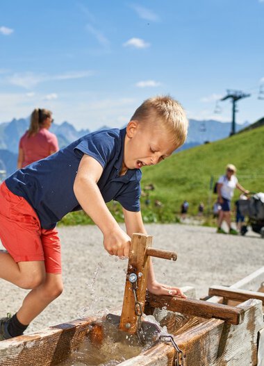 Boy playing with water fountain outdoors in the mountains, jumping happily. | © Oberstdorf · Kleinwalsertal Bergbahnen Photographer: Christian Seitz