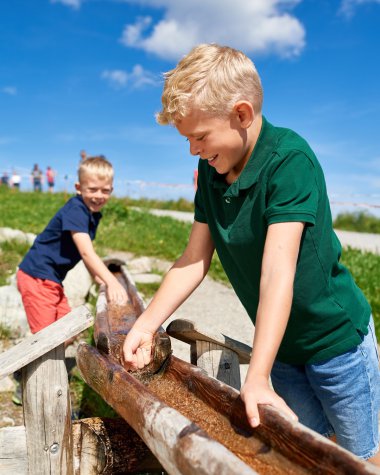 Two children drawing water from a natural water source, surrounded by greenery and a blue sky with clouds. | © Oberstdorf · Kleinwalsertal Bergbahnen Photographer: Christian Seitz