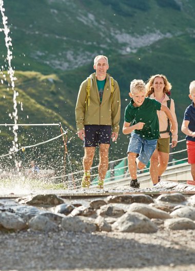 Family with two children barefoot walking through a water feature in the mountains, with green mountain forest and a cable car in the background. | © Oberstdorf · Kleinwalsertal Bergbahnen Photographer: Christian Seitz