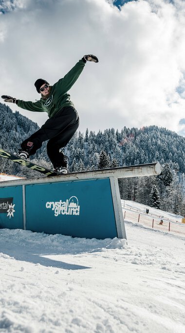 Skater performing a rail stand on a snowboard in a snowy ski area with snow-covered mountains and clear sky. | © Kleinwalsertal Tourismus eGen • Crystal Ground • Photographer: Stefan Eigner