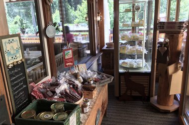 Interior of a small farm shop with products on a wooden shelf and in a refrigerated display, with double doors in the background, lots of natural light. | © Kleinwalsertal Tourismus eGen • Walser Buura Café Baad Grund