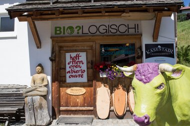Entrance to Kasestadl farm shop in Hirschegg, with a green roof, wooden sign, a butter sculpture, and a bench, under a blue sky. | © Kleinwalsertal Tourismus eGen • Käsestadl