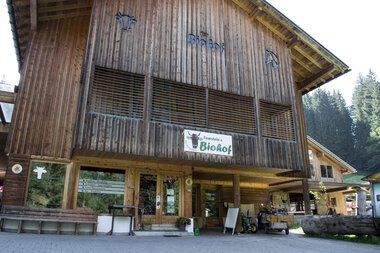 Wooden farm shop with organic farm sign in Kleinwalsertal. Front view with entrance, bicycle, and benches. | © Kleinwalsertal Tourismus eGen • Biohof Feurerstein