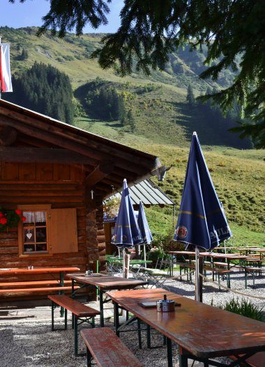 Mountain restaurant in the Alps with several tables and black umbrellas outdoors, surrounded by green meadows and mountains. | © Kleinwalsertal Tourismus eGen • Mittelalp