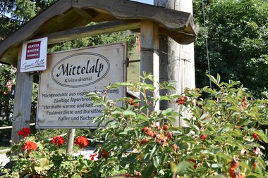 Sign for the GenussRegion Mittelalp in Kleinwalsertal, surrounded by red flowers and green plants. Wooden posts and trees in the background. | © Kleinwalsertal Tourismus eGen • Mittelalp
