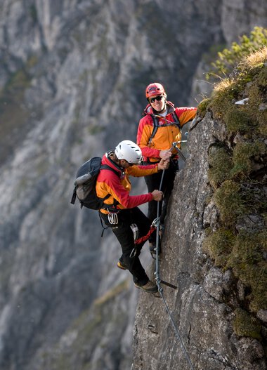 Two climbers on a via ferrata in the mountains, securing themselves to a narrow rock ledge. | © Kleinwalsertal Tourismus eGen • Bergschule