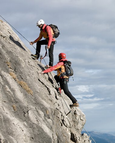 Two climbers with gear and helmets ascending a steep rock wall on a climbing route in Kleinwalsertal. | © Kleinwalsertal Tourismus eGen • Bergschule