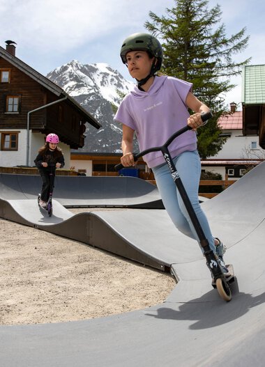 Two children, a girl and a boy, riding scooters on a skate park in the mountains with wooden and stone houses in the background. | © Kleinwalsertal Tourismus eGen • Photographer: Frank Drechsel