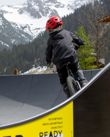 Two children riding bicycles on a pump track in a mountainous area with snow-capped peaks in the background. | © Kleinwalsertal Tourismus eGen • Photographer: Frank Drechsel