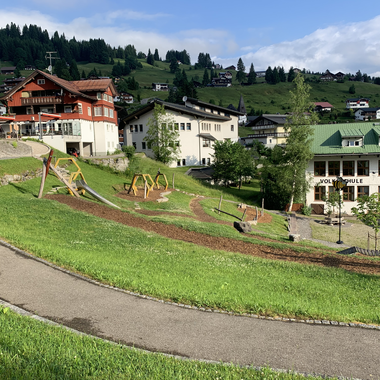 View of a playground in Hirschegg with slide, swings, and paths, surrounded by green grass and mountains in the background. | © Kleinwalsertal Tourismus eGen