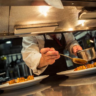 A chef carefully pouring sauce over prepared dishes in a professional kitchen. | © Kleinwalsertal Tourismus eGen • Photographer: Oliver Farys