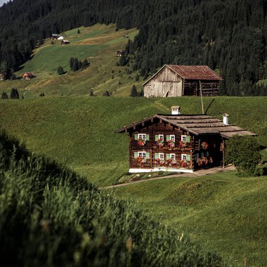 A traditional Walser house amidst green hills with mountains in the background, set in a scenic landscape. | © Kleinwalsertal Tourismus eGen • Photographer: Oliver Farys