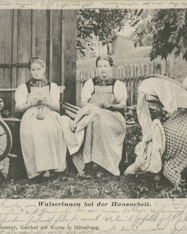 Four women in traditional costume sitting outdoors in a garden, working on various crafts. | © Kleinwalsertal Tourismus eGen